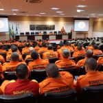 Bombeiros militares de uniforme laranja participam de uma atividade de treinamento estratégico ao ar livre.