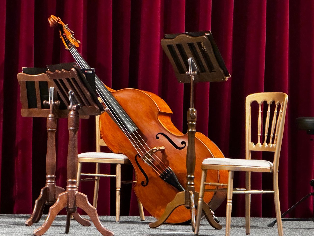 Palco vazio preparado para um concerto, com um violoncelo e estantes de partituras.
